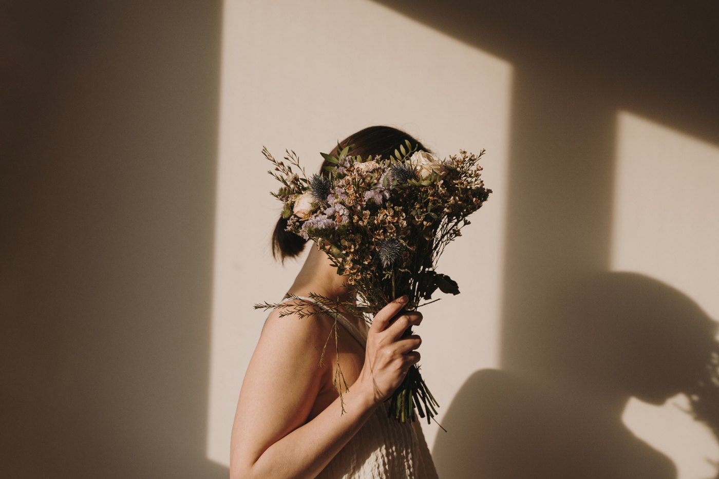 A woman holding a bunch of dried flowers in front of her face, standing in warm window light